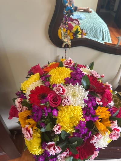 A large bouquet of flowers sits on a table in a residence in Río Oro, Costa Rica, at coordinates 9.945, -84.201. The bouquet is composed of various flowers, including red, yellow, white, purple, and pink chrysanthemums, red gerbera daisies, pink roses, and sunflowers. The bouquet is arranged in a circular pattern and is surrounded by green leaves. The flowers are all in bloom and appear to be fresh. A mirror in the background reflects a bed with a green and white patterned comforter and a figure laying on it. The image is taken from an angled perspective, looking down at the bouquet. The focus is on the flowers, and the background is blurry.