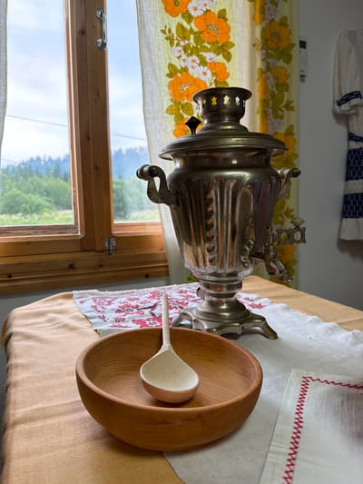 A close-up, eye-level shot captures a still life arrangement on a table with a window and floral curtains in the background. The table is covered with an orange cloth, a white cloth with red embroidery, and a piece of patterned fabric. In the foreground, a wooden bowl contains a wooden ladle. Behind the bowl, a silver samovar stands on a decorative base, featuring a spigot and handles. The window behind the samovar reveals a verdant, hilly landscape with trees. The curtains, patterned with orange and yellow flowers, hang to the right of the window. On the wall to the far right, a piece of embroidered clothing is visible. The scene suggests an indoor setting in Ust'-Kabyrza, Russia, possibly a home or a traditional craft shop, with elements of domesticity and regional culture.