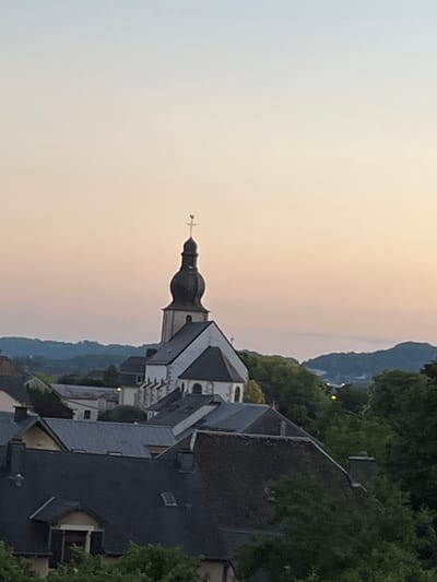 This is a photograph of the town of Mondercange, Luxembourg, taken from an elevated viewpoint, likely a hillside overlooking the town. The image captures a scene at dusk, with a soft, gradient sky transitioning from pale blue at the top to a peachy pink near the horizon.
Dominating the central area of the image is a church with a tall, dark-colored steeple topped with a golden cross. The church itself is white with dark grey roofs, and appears to be an older architectural style. Surrounding the church are numerous buildings with steeply pitched roofs, characteristic of European towns. These buildings are predominantly dark grey and brown, suggesting they are covered in slate or similar roofing materials. Some buildings exhibit dormer windows and chimneys.
The foreground is filled with the rooftops of residential or commercial buildings, mostly in dark grey tones. Lush green trees are interspersed throughout the town and in the surrounding hills, indicating a setting with significant vegetation. In the background, rolling hills are visible, fading into the hazy evening light. The overall impression is one of a tranquil, picturesque town settling into the evening. There are no people visible in the image.