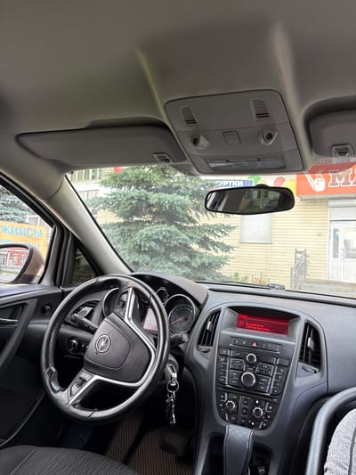 The image shows the interior of a car, likely a modern hatchback or sedan, with the perspective looking towards the front windshield. The driver's side of the car is visible, including the steering wheel, dashboard, and center console. The steering wheel is black with silver accents and features the Opel logo in the center. Buttons for audio and cruise control are visible on the steering wheel.
The dashboard includes an instrument cluster with a speedometer and tachometer, the latter showing a redline at approximately 6000 RPM. To the right of the instrument cluster is the car's infotainment system, which has a red digital display showing "15:01" and text in Russian indicating a connected AUX device. Below the display are various buttons and knobs for controlling the radio, CD player, climate, and other vehicle functions. The gear shifter is visible in the center console, topped with a quilted leather-like cover.
The view through the windshield shows an outdoor scene. There are large evergreen trees, suggesting a location with significant vegetation. A building with yellow siding and a red sign with Cyrillic lettering (partially visible as "УРТКИ") is behind the trees. Another sign with "ДЖИНСЫ" (Jeans) in large yellow letters is visible on the left side. The overall environment suggests an urban or suburban setting. Based on the provided information, the location is the city of Rezh, Russia. There are no people visible in the image.