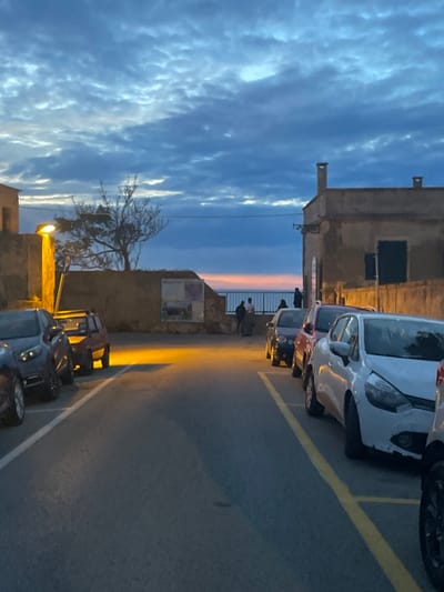 The image shows a street scene in Portoferraio, Italy, taken at dusk. The camera is facing down the street. The street is lined with parked cars, including a white car in the foreground. There are several people standing by a wall in the distance, looking out at the sea.  The sky is blue with pink clouds. The image was taken at latitude 42.817 and longitude 10.332. The street is paved with asphalt, and the street lights are on.
