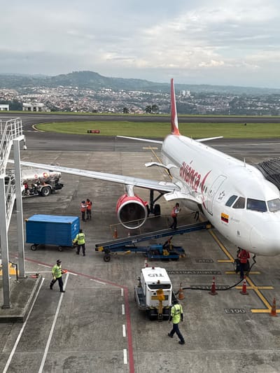 An Avianca Airbus A320 aircraft, predominantly white with red tail markings and engine nacelles, is stationary on a concrete tarmac at an airport in the city of AMCO, Area Metropolitana Centro Occidente, Colombia. The Colombian flag is visible on the aircraft's forward fuselage. Ground operations are actively underway. A white Terpel fuel truck is positioned beneath the aircraft's left wing, while a blue baggage container on wheels is being pushed by a ground crew member wearing a high-visibility yellow-green vest. A blue conveyor belt loader is connected to a cargo hold opening beneath the aircraft, with a crew member standing on it. A white ground power unit, labeled "GT1," is located near the aircraft's nose, where fueling hoses are connected and demarcated by orange safety cones. In total, approximately seven ground crew personnel, attired in high-visibility yellow-green or orange vests, are present around the aircraft, performing various tasks. The tarmac displays painted markings including 'AT42', 'A320/AT72', 'E190', and red and white boundary lines. In the background, a curved asphalt runway or taxiway with markings '0 26 08' is visible, separated from the tarmac by a strip of green grass. Beyond the airport facilities, a dense urban area is visible across undulating hills under an overcast sky.