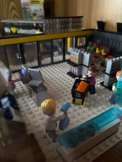 This image shows a Lego diorama of a supermarket. The diorama is set in Delbrück, Germany, as indicated by the text on a sign in the background which reads "Wir haben Lebensmittel" (We have groceries) and the Edeka logo on a shopping cart. Several Lego minifigures are present. One minifigure, appearing to be an elderly woman, stands at a checkout counter with a Lego computer and scanner. Another minifigure, a child with blonde hair, pushes a shopping cart containing a blue object and what appears to be a loaf of bread and another orange item. A third minifigure, a young woman with brown hair, pushes a shopping cart with a loaf of orange bread inside, also bearing the Edeka logo. A partially visible minifigure in a green shirt stands to the right. The supermarket features shelves stocked with Lego products, a checkout area with a Lego register, and a display case with blue items. The floor is made of white Lego plates. The overall scene depicts a typical day at a supermarket.