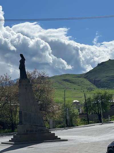 The image shows a monument in Sisian, Armenia. The monument consists of a statue atop a stone pedestal, with a plaque visible on the side. Trees surround the monument, and a road with steps leading up to the monument is visible. In the background, there is a grassy hill and a sky with puffy white clouds. Power lines are also present in the frame. A portion of a dark-colored vehicle is visible in the top right corner of the image.