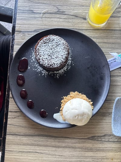 A top-down view of a black plate on a wooden table shows a chocolate lava cake topped with powdered sugar alongside a scoop of vanilla ice cream on a bed of crushed graham crackers, and five small droplets of chocolate sauce arranged in a semi-circle. A glass of orange juice sits to the left, and a sugar packet is visible at the top. Based on the image details, it is not possible to determine the specific location within Veracruz, Mexico.