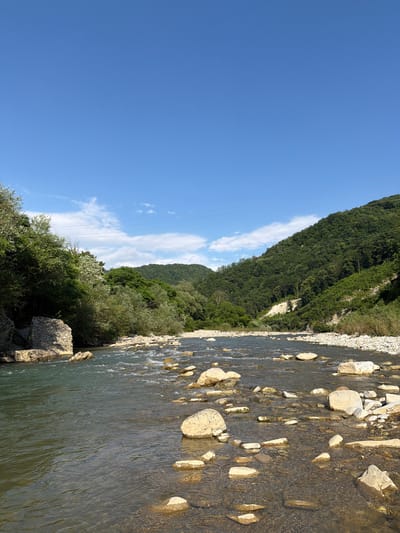 The image depicts a natural landscape featuring a river flowing through a rocky riverbed, bordered by lush green hills and trees under a clear blue sky. The river in the foreground has calm, clear water, revealing numerous smooth, rounded stones on its bed. The riverbanks are lined with more stones and vegetation. In the background, the hills are densely covered with trees, suggesting a forest. The sky is predominantly blue with a few scattered white clouds and power lines are visible across the upper portion of the image. There are no people or animals present in the scene. The location is identified as Krasnoye, Russia.