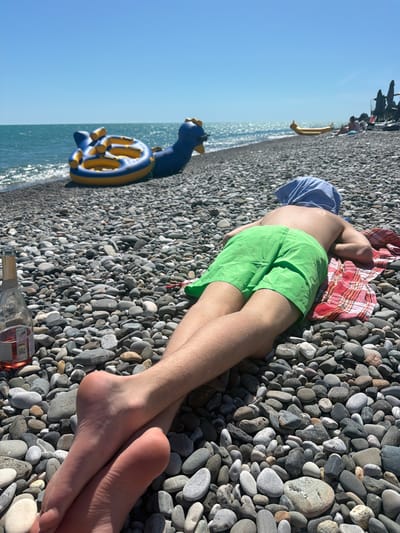 A person is lying on a pebble beach in Amzara, Abkhazia. They are wearing green shorts and a blue head covering. Their feet are crossed. A bottle of rosé wine is placed on the beach to the left of the frame. An inflatable toy shaped like a dinosaur is in the water behind them, and a yellow inflatable raft is further down the beach. The sea is visible under a clear blue sky. Several beach umbrellas and chairs are in the background.