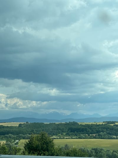 The image depicts a panoramic view of the Bulgarian countryside under a dramatic sky. In the foreground, a blurred metal railing runs horizontally across the bottom, suggesting the viewpoint is from a vehicle. Beyond the railing, the landscape unfolds with rolling hills covered in lush green trees and fields of golden-yellow crops. A winding road with a small building is visible in the middle ground. In the distance, a range of mountains with snow-capped peaks stands against a backdrop of a heavy, cloudy sky. The lighting suggests it is daytime, possibly late afternoon or early evening, with the clouds casting a diffused, cool light over the scene. The location is identified as being near Vakarel, Bulgaria, which is a village situated in the Sofia Province known for its natural beauty and proximity to mountains.