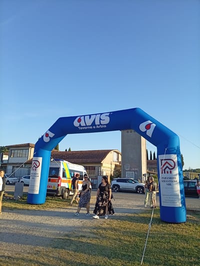 An inflatable blue archway branded with "AVIS" and "Taverne e Arbia" stands under a clear blue sky. The archway is sponsored by "PARASILITI SEBASTIANO SRL," a construction company. Behind the archway, an ambulance with red and yellow stripes is parked, and several people are walking or standing around. Buildings with tiled roofs are visible in the background, along with trees and power lines. The ground is covered with gravel and grass. The scene appears to be an outdoor event or gathering in the town of Arbia, Italy.