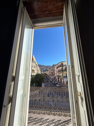 The image shows an open doorway with a view of a street in Maddaloni, Italy. The doorway is framed by white painted wood. Through the doorway, a street lined with buildings is visible. The buildings are colorful and appear to be old. There are cars parked along the street and a few trees. The sky is clear and blue. The balcony railing in the foreground is made of wrought iron with an ornate pattern. The sun casts long shadows on the balcony floor. A brass door handle is visible on the right door.