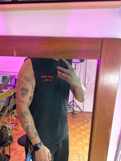 A mirror selfie reveals a person in a black t-shirt with red lettering, partially obscuring their face. Visible skin displays multiple tattoos. The background includes a room with a wooden floor, visible cords, a white wall, and what appears to be a plant in front of a computer screen. The mirror has a wooden frame, with pink lighting reflected on the adjacent wall.