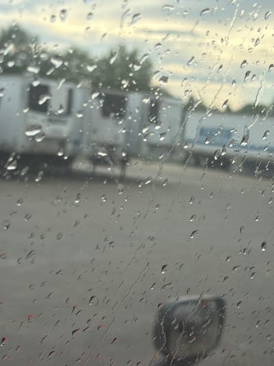 The image shows a view through a rain-streaked window, likely from inside a vehicle. In the background, several white semi-trailers are parked, suggesting a commercial or industrial area. The sky above appears overcast with hints of pale yellow light, possibly indicating dawn or dusk. The foreground is dominated by numerous raindrops on the glass, distorting the view of the scene beyond. The ground appears to be a paved surface, possibly asphalt. A blurred reflection of what looks like a vehicle's side mirror can be seen in the lower right portion of the image. The presence of semi-trailers suggests the location could be a truck stop, distribution center, or a similar facility in or around Murfreesboro, Tennessee. There are no clearly discernible people or specific actions taking place.