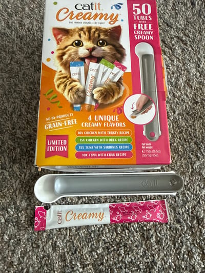 An overhead shot displays a box of Catit Creamy cat treats, a cat treat tube, and a grey and white cat treat dispenser, all situated on a grey carpeted floor. The box is predominantly orange and white and features an image of a kitten. Text on the box reads "Catit Creamy," "The Famous Lickable Cat Treat," "50 Tubes," "With Free Creamy Spoon," "No By-Products," "Grain-Free," "4 Unique Creamy Flavors," and a list of flavors with quantities: "10x Chicken with Turkey Recipe," "15x Chicken with Duck Recipe," "15x Tuna with Sardines Recipe," and "10x Tuna with Crab Recipe." A pink banner on the box indicates "Limited Edition." The treat tube is white with pink crabs and fish, and the text "Catit. Creamy." The dispenser is grey and white with the "Catit" logo. The image was taken in Girard, United States of America.