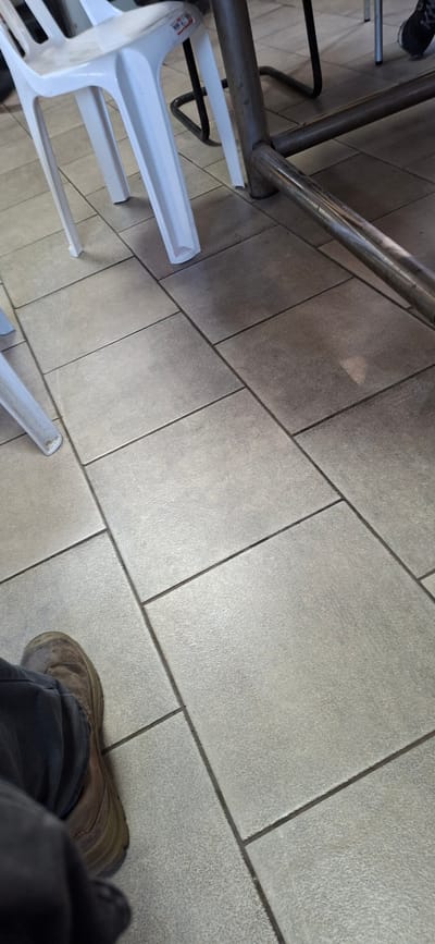 The image shows a floor tiled with large, light grey rectangular tiles. A person's foot, clad in a brown, worn shoe and dark pants, is visible in the lower left quadrant, resting on the tiles. In the upper left, a white plastic chair with a curved back and four legs is partially visible, its legs also resting on the tiled floor. Behind the chair, a metallic structure, possibly part of a table or a railing, is present, composed of cylindrical metal tubes. The environment appears to be indoors, potentially a restaurant or a public space, with natural light illuminating the scene. The location is identified as Jezreel Valley Regional Council, Israel.