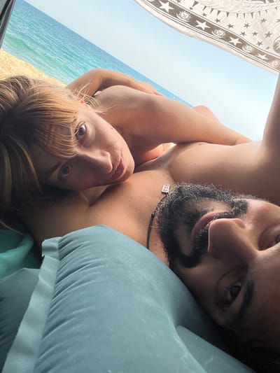 Two individuals, a man and a woman, are lying down outdoors. The man, in the foreground, has dark hair and a beard and is looking towards the camera. He is wearing a necklace with a tag that says "RAW." The woman, positioned behind him, has lighter hair and is looking towards the camera with her arm around him. They appear to be on a beach, with the blue ocean and sand visible in the background. A patterned fabric, possibly a beach towel or canopy, is visible at the top of the frame. The image suggests a casual and intimate moment between the two. The location is identified as Sant Pol de Mar, Spain.