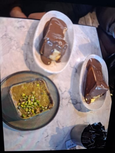 The image depicts a tabletop scene within an establishment in Selyanin, Bulgaria. The surface is a light-colored, possibly marble-patterned, table. Three dessert items are prominently displayed. On the left, a square-shaped dessert, possibly Kunefe or similar, with a greenish-yellow top layer covered in chopped pistachios, is presented in a rectangular dish placed on a dark-rimmed plate. To the right, two oval-shaped white dishes each contain a portion of light-colored cake or pastry completely coated in dark brown chocolate sauce or ganache. Below the rightmost chocolate dessert, a light grey cylindrical container is filled with numerous individually wrapped black sachets, likely sugar packets. A small white rectangular card displaying the text "RODIN" and a QR code is visible near this container. Portions of human limbs are partially visible in the upper and right edges of the frame, suggesting individuals are seated around the table. No direct actions or interactions are visible, but the setup indicates a dining or dessert-consuming activity.