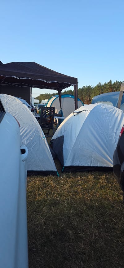 The image depicts a campsite in Neustadt-Glewe, Germany, on a clear day. Several tents are set up on a grassy area, with a dark-colored canopy providing shade. In the foreground, the side of a white car is visible, partially obscuring a light-colored tent. Further back, more tents and what appear to be caravans or RVs are present, with a line of trees forming the backdrop. A black folding chair is positioned near a table under the canopy. The ground is covered with dry grass. There are no people visible in the frame.