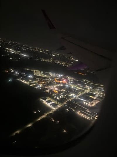 The image, taken from an airplane window at night, depicts a cityscape illuminated by countless lights. The view looks down on a grid-like arrangement of buildings and streets, suggesting an urban or suburban area. The lights create a shimmering effect, with some appearing as bright, distinct points and others as diffused glows from illuminated windows or street lamps.

In the foreground, the edge of an airplane wing or fuselage is visible, along with a portion of the airplane window. A purple flag or banner is attached to the wing, and another similar object with red and white stripes is visible further along the wing.

The scene is set at night, with the darkness of the sky contrasting sharply with the man-made lights of the city below. There are no visible people, but the illuminated structures imply human habitation and activity. The weather appears clear, allowing for an unobstructed view of the city lights. The location is identified as Villafranca di Verona, Italy. The overall mood evoked is one of wonder and observation, looking down upon the sleeping city from above.