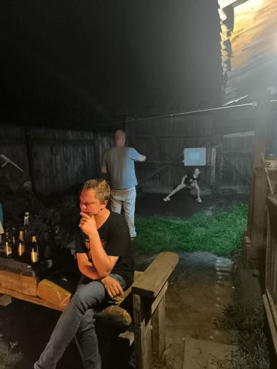 A dimly lit outdoor scene, possibly in or around a rural area like Кашино, Russia, features several individuals and objects. In the foreground, a man with blonde hair, wearing a black t-shirt and blue jeans, is seated on a wooden bench, appearing to be in thought or resting his chin on his hand. Several bottles, likely alcoholic beverages, are lined up on a wooden surface next to him. In the middle ground, a bald man in a grey t-shirt and light-colored pants stands facing away from the camera, gesturing towards something off-frame. Further back and slightly to the right, a woman in a dark t-shirt and shorts is captured in a dynamic pose, possibly exercising or dancing, with her legs spread wide. The background consists of wooden structures and fencing, with a patch of green grass in the immediate foreground and more grass and darker foliage further into the scene. The lighting suggests it is nighttime, with artificial light sources illuminating parts of the area.