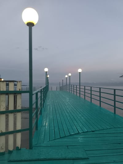 A wide wooden pier, painted a vibrant teal, stretches into the distance under a pale, overcast sky. The pier's planks are laid lengthwise and appear slightly wet. Along the right side of the pier, a dark teal railing follows its course. Scattered at intervals along the pier are spherical, lit lamps mounted on tall, slender poles. In the background, the calm, gray water of a lake or sea is visible, with a distant, hazy shoreline. To the left, a white wooden fence with vertical slats is partially visible, also bordering the pier. The scene is set at dusk or dawn, indicated by the subdued natural light and the illuminated lamps. The location is identified as the city of Bayet, Kyrgyzstan. No people are present in the image.