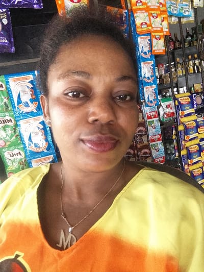 This image depicts a close-up portrait of a single adult female in what appears to be a small retail shop or provision store in Usiefrun, Nigeria. The woman, of apparent African descent, is centered in the frame, looking directly at the camera with a slight, closed-lip smile. She has dark, short, curly hair. Her ears each feature a small, light-colored stud earring. She wears a silver-colored chain necklace with a pendant shaped like the letter "M." Her attire consists of a garment with a V-neck, displaying a bright yellow upper section that transitions into an orange and darker orange tie-dye pattern below the chest line.
The background reveals an interior retail setting with various packaged goods and bottled items displayed on shelves and hanging from hooks. To the woman's left (viewer's right), several green and blue sachets are visible, identifying products such as "Milo" and "Combell" milk powder. To her right (viewer's left), a denser display includes rows of yellow and orange packaged items (e.g., "Cheeseballs" biscuits), numerous smaller sachets of various food products (e.g., "OYINO" seasoning, "COMFORT" sweets), and a shelf stacked with bottles of what appear to be spirits or other beverages. The products suggest a typical local grocery or convenience store environment. The lighting is bright, illuminating the woman's face and the merchandise.