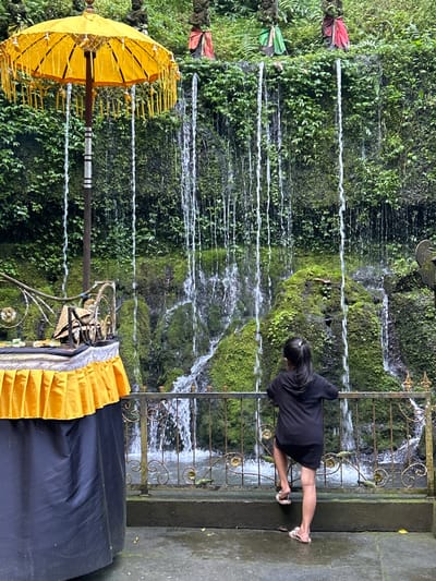 A young girl in a black hoodie dress and flip-flops leans on a decorative metal fence, looking at a moss-covered rock face with multiple thin waterfalls cascading down it. In the background, three stone figures adorned with colorful cloths stand among lush green foliage atop the rock face. To the left, a large, ornate yellow umbrella with decorative tassels stands next to a table draped with a black cloth and a yellow pleated fabric skirt. The ground is wet, suggesting recent rain or spray from the waterfalls. The scene appears to be in an outdoor, natural setting, possibly a temple or sacred site, given the presence of statues and offerings. This location is identified as the Tirta Empul Temple in Bangli, Indonesia.