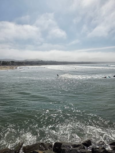 The image depicts a coastal scene in Dana Point, California, United States of America, on a hazy day. The foreground is dominated by dark, wet rocks along the shoreline, with foamy waves breaking against them. The ocean stretches into the midground and background, appearing choppy with sunlight glinting off the surface. Several individuals are visible in the water, some on surfboards, with one person actively surfing a wave. The shoreline in the distance is lined with a sandy beach populated by numerous people. Further inland, a gentle, tree-covered slope rises, with buildings and palm trees visible on its crest. The sky above is a pale blue, filled with scattered clouds, some of which are low-lying and appear as fog banks over the distant landmass.