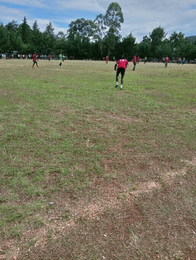 An outdoor football (soccer) match is in progress within Lwandeti ward, Kenya. The foreground and mid-ground consist of a large, partially dry grassy field interspersed with patches of exposed soil and dirt, with faint white field markings visible on the ground. A male player, wearing a red jersey with the number "11" on the back, black shorts, and blue and white cleats, is prominently featured in the foreground from a rear perspective, bent forward in a posture suggesting active movement. Approximately ten additional male players are distributed across the field, with some wearing red jerseys and others wearing green jerseys, all engaged in the football match. The background is dominated by a dense line of trees, including coniferous and tall deciduous species. A crowd of spectators is visible along the tree line. The sky above is partly cloudy.