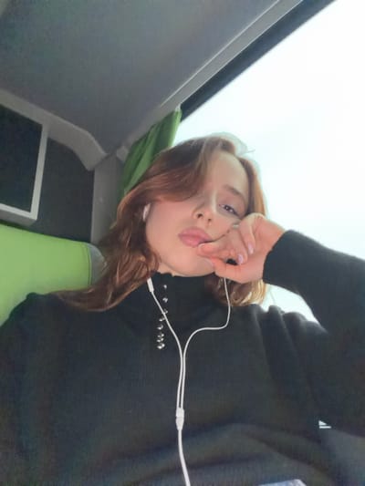 A young female, with shoulder-length reddish-brown hair parted on the left, is depicted from the chest up, taking a selfie inside what appears to be a passenger vehicle, likely a bus or coach, in Moineville, France. She wears a black ribbed turtleneck sweater with silver-colored ring fasteners on the collar and white wired earbuds in both ears, the cable extending down her chest. Her left hand is raised, with fingers slightly curled, thumb and index finger touching her lower lip area. A ring is visible on her left ring finger, and her fingernails are painted a light shade. Her lips are slightly parted, revealing a portion of her tongue, and she is looking directly at the camera. In the background, to the left, a portion of a green upholstered seat and a rectangular screen are visible. To the right, a large window shows a bright, overcast sky. A green curtain or fabric element is partially visible near the window's top left edge. The overhead ceiling panel is gray.