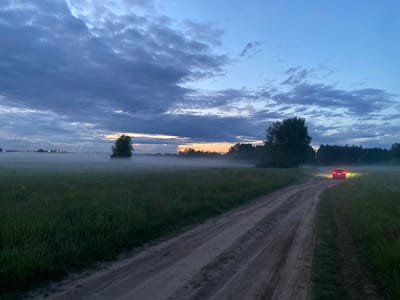The image depicts a rural road at dusk, likely in Yablonnoe, Russia. A dirt road curves through a field of tall grass, leading towards a stand of trees in the distance. A car with its headlights and taillights illuminated is visible on the road, approaching the viewer. A light mist hovers over the field, particularly in the background, adding to the atmospheric quality of the scene. The sky is a gradient of deep blue and grey clouds, with a faint glow of sunset on the horizon. The overall impression is one of a quiet, serene evening in the countryside.