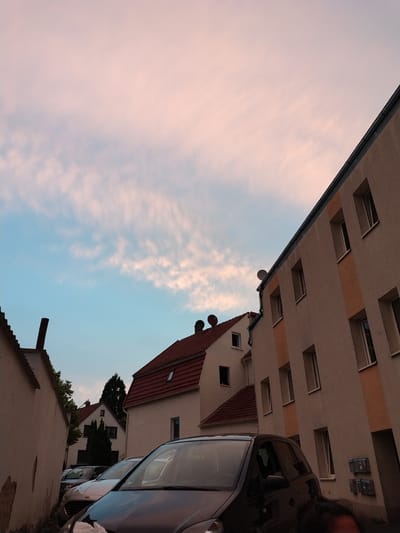 The image shows a street scene in Herford, Germany, with buildings and cars. The sky is visible in the background, with soft pink and blue clouds. The buildings are multi-story structures with pitched roofs and windows. There are several cars parked on the street. The overall impression is of a quiet, residential area.