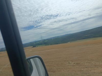 The image, taken from a vehicle possibly traveling through Nohn, Germany, shows a rural landscape under a cloudy sky. In the foreground, a dark-colored pillar, likely part of the vehicle's interior structure, runs vertically along the left side of the frame. A wing mirror is visible in the lower right portion of the image, reflecting a portion of the sky and possibly the vehicle's interior.

The mid-ground features a vast, dry, golden-brown field, suggesting a harvested crop such as wheat or barley. The field stretches horizontally across the image, with some texture and slight undulations. In the distance, a dense, green forest covers rolling hills, creating a stark contrast with the harvested field. A tall, latticed electricity pylon stands prominently against the sky in the far-left mid-distance, with power lines extending from it. The sky is filled with a complex pattern of white and gray clouds, with hints of blue sky peeking through in the upper left. The overall impression is one of a serene, agricultural countryside.