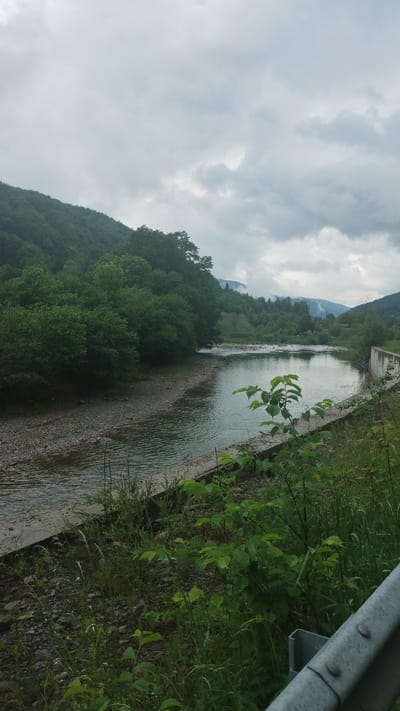 The image shows a river flowing through a landscape in Trostianets, Ukraine. The riverbanks are lined with lush green vegetation and trees, and a mountain range is visible in the background. A concrete barrier and metal guardrail run along the riverbank. The sky is overcast with heavy clouds.