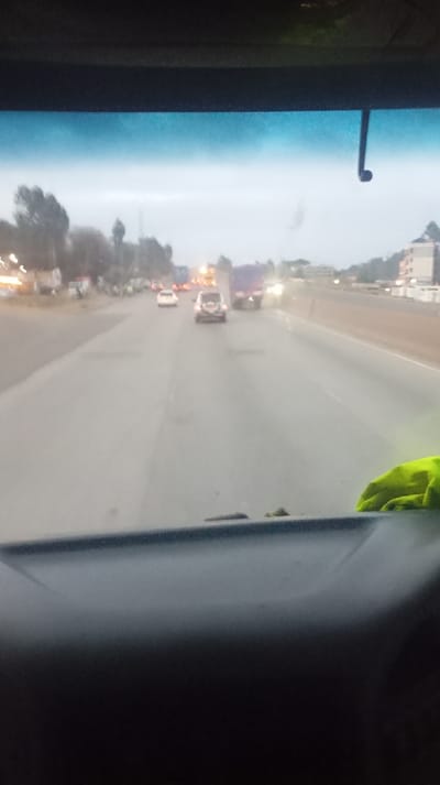 A road scene viewed from inside a vehicle, likely a car or truck. Several vehicles are on the road ahead, including a truck. The road appears to be multi-lane and is relatively straight. Buildings are visible on the right side of the road. A green cloth is visible on the right side of the vehicle. The image is somewhat blurry.
