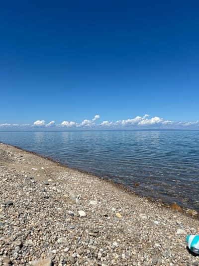 The image depicts a serene beach scene under a clear blue sky, with a vast expanse of calm water stretching to the horizon. The foreground is a pebbly beach, with smooth, varied-colored stones leading down to the water's edge. The water itself is shallow and clear, revealing more stones beneath the surface. A faint line of clouds dots the distant horizon, separating the water from what appears to be a landmass. A portion of a blue and white striped object, possibly a beach mat or float, is visible in the lower right corner of the frame, suggesting human presence or recent activity. The overall impression is one of tranquility and natural beauty. The scene is identified as being in the city of Tolbazikha, Russia.