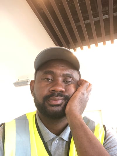 The image depicts an adult male situated indoors, presenting a direct gaze into the camera. He possesses a dark skin tone, a full dark beard, and short dark hair. He wears a grey baseball cap, a grey collared polo shirt, and a bright yellow high-visibility vest accented with grey reflective stripes. His left hand is positioned against the left side of his face, appearing to support his head. The immediate background features a uniformly light-colored wall and ceiling. An off-white rectangular fixture, possibly an air conditioning unit, is visible on the upper left portion of the wall. The upper right portion of the image reveals a ceiling structure composed of multiple parallel dark slats or beams. The environment is brightly illuminated by diffuse light. No specific visual elements within the image content definitively identify the location as being within the city of Hanak, Saudi Arabia.