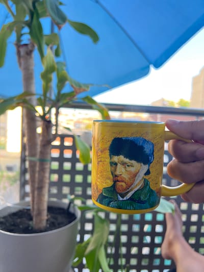 A hand holds a yellow mug featuring a painting of a man's face, possibly Vincent van Gogh's "Self-Portrait with Bandaged Ear". The mug is held horizontally and tilted downwards. In the background, a blue umbrella is partially visible to the left, and a potted plant with green leaves is in the foreground to the right. Beyond the plant, a black lattice-like structure and a glimpse of a building can be seen. The lighting suggests it is daytime, with sunlight casting bright reflections and highlights. The scene appears to be outdoors, possibly on a balcony or patio, with the background suggesting an urban or suburban environment in Mollet del Vallès, Spain.