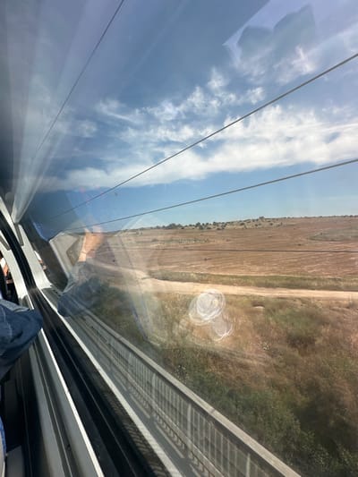 The image shows a view from inside the Modi'in-Maccabim-Re'ut, Israel high-speed train. Through the window, a harvested field and a dirt road can be seen on the right, while the sky with clouds is reflected on the left. There are several train power lines. Part of the train interior is visible at the bottom, including an armrest and a leg with a blue jean. There is also a water droplet stain on the window.