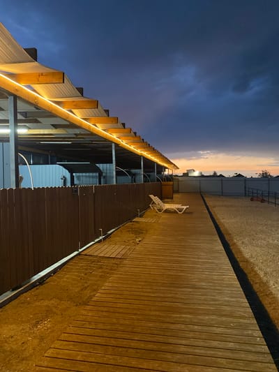 The image depicts an outdoor scene at dusk, possibly a recreational area in Izberbash, Russia. A wooden boardwalk extends from the foreground to the mid-ground, running parallel to a brown wooden fence. The boardwalk is illuminated by warm-toned lights installed along the edge of an overhang structure. To the left of the boardwalk, beneath the overhang, are some structures and what appear to be shower stalls or similar amenities. In the mid-ground, on the sandy area to the right of the boardwalk, two white lounge chairs are placed. The background shows a fence surrounding a sandy area, and beyond that, the silhouettes of buildings and trees against a twilight sky, with hints of orange and pink from a setting sun. The sky is a deep blue with dark clouds. A small red garden roller is visible in the far right of the image. No people are present in the scene.