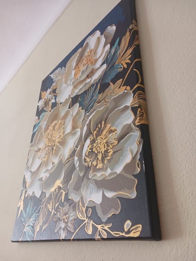 A painting of three large, light-colored flowers with gold accents is hanging on a cream-colored wall. The painting is angled, with the top left corner higher than the bottom right. The flowers have detailed gold centers and highlights on their petals. Surrounding the flowers are gold leaves and some blue-green foliage. The background of the painting is a dark navy blue. The wall appears to be textured. The scene is indoors, suggesting it is in a room within a building. There are no people visible in the image.