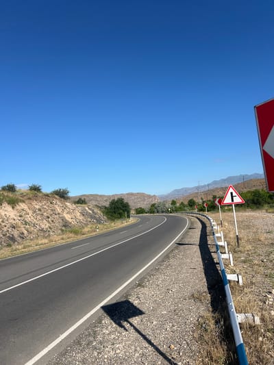 The image depicts a clear, blue sky over a rural, mountainous landscape in Malishka, Armenia. A paved asphalt road curves gently to the left, with white lane markings visible. On the right side of the road, a metal guardrail separates the road from a dry, grassy embankment littered with gravel and stones. Several road signs are present: a large, red and white directional sign is partially visible on the far right, and further down the road, a triangular sign with a black symbol indicating a T-junction is mounted on a white pole. Another smaller, red and white directional sign is also visible. Dry, golden grass and sparse bushes dot the landscape, with trees and hills in the background. Power lines and poles are also present in the distance. The sun casts a distinct shadow of a square object onto the road.