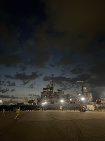 At dusk in Verkhnyaya Stavropol'ka, Russia, a large ship, identified by the number "105" on its hull, is moored at a quay. The ship's superstructure is visible, with various antennas and equipment. The sky is dark with scattered clouds, illuminated by artificial lights. In the foreground, a paved area is occupied by a few pedestrians and a three-wheeled vehicle. One person, wearing a yellow shirt, is walking across the paved area. Another person, in lighter clothing, is walking further to the left. The three-wheeled vehicle, with an orange canopy, is carrying passengers and is moving away from the camera. Streetlights line the quay, casting pools of light on the ground. The overall scene depicts a harbor area at the end of the day, with the imposing presence of the ship dominating the background.