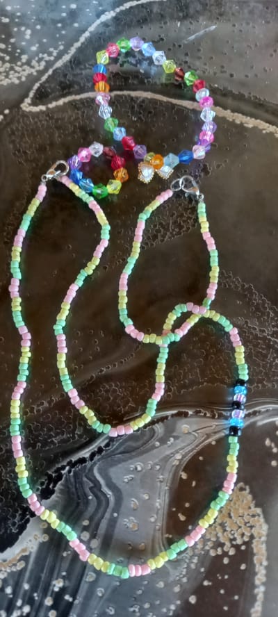 This is a close-up, top-down shot of two beaded necklaces laid out on a dark, marbled surface. The necklace on the left is made of faceted, brightly colored beads in red, orange, yellow, green, blue, purple, and clear. It also features a small, metallic charm shaped like a butterfly with white enamel. The necklace on the right is made of smaller, matte beads in pastel shades of pink, yellow, and light green, forming a repeating pattern. Towards the top right of this necklace, there are a few black, blue, and white beads, potentially forming a clasp or accent. The surface appears to be a table or countertop with a swirling, abstract pattern of black and white, possibly representing stone or resin. There are no people visible, and the scene suggests a still-life arrangement of jewelry. The lighting is even, indicating an indoor setting, possibly a crafting area or a display. There is no visible text in the image.