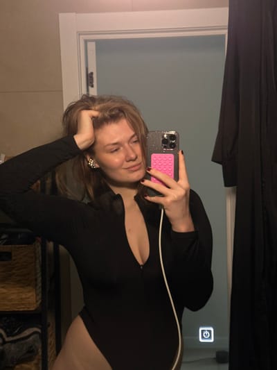 A young adult female is depicted in a mirror selfie, taken indoors in Бакча-Сарай, Russia. She holds a dark-colored smartphone, equipped with a sparkly case and a pink pop-socket, in her right hand. A white charging cable is connected to the phone. Her left hand is positioned on the top of her head, lightly touching her light brown hair. She is wearing a long-sleeved black bodysuit with a partially unzipped front, revealing cleavage. A prominent, light-colored earring is visible on her left ear. Her fingernails on the right hand are painted a dark red or maroon color. The background, reflected in the mirror, includes a light-colored wall, a white door frame with a closed door revealing a light blue/gray room, a shelving unit with woven baskets and textiles on the left, and dark fabric hanging on the right. A glowing white power icon is visible on the lower right corner of the mirror surface.