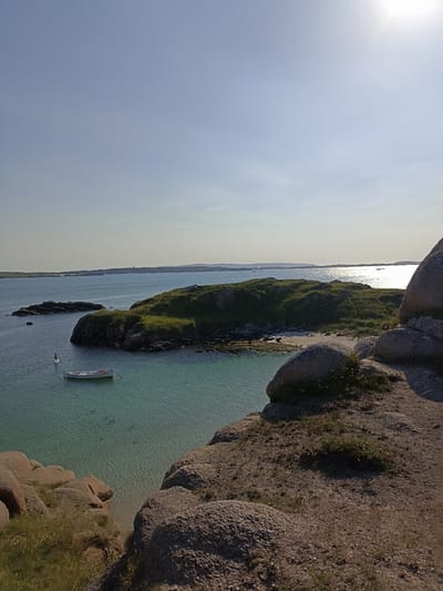 The image depicts a serene coastal landscape. In the foreground, large, rough granite boulders are prominent, with sparse grass growing between them. A clear, turquoise body of water occupies the middle ground, revealing the sandy seabed below. A small, white boat with orange trim floats on the water, anchored by a rope. Further in the distance, a green, grassy island with rocky outcrops rises from the sea. A single person on a paddleboard is visible in the water between the island and the foreground. The background shows a calm sea stretching to the horizon, with distant islands or landmasses under a clear, bright sky. The sun is high and slightly to the right, casting a bright glare on the water. The overall impression is one of a peaceful, natural setting on a sunny day. The location is identified as Carnboy, Ireland.