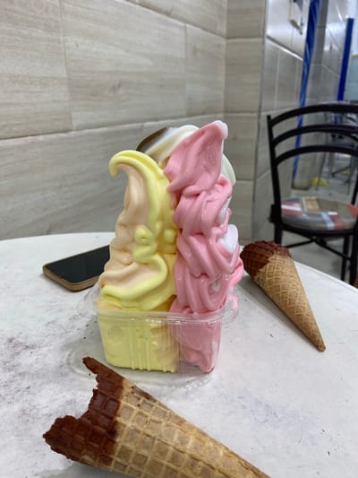 The image depicts a close-up of a triple-scoop soft-serve ice cream in a clear plastic container. The ice cream has three distinct flavors and colors: pale yellow, pink, and white, with swirls of chocolate on top. Two waffle cones are present, one partially dipped in chocolate and lying in the foreground, with a bite taken out of it. Another plain waffle cone is positioned to the right of the ice cream. A smartphone is visible on the left side of the table, near the ice cream. The background shows a tiled wall and a black chair, suggesting an indoor setting, possibly a shop or cafe. The scene is set on a textured white table surface. The lighting is natural and appears to be from a window or overhead source, casting soft shadows. The image does not contain any information that would suggest a specific location in Sidi Chahmi, Algeria.