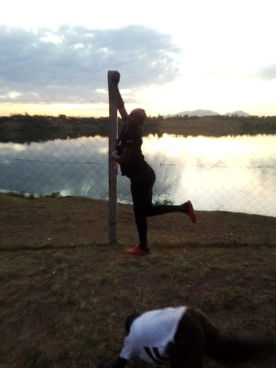 The image displays an outdoor scene in Central Kasipul ward, Kenya, during a low-light period, likely dawn or dusk. In the mid-ground, a person, silhouetted against the bright horizon, stands next to and holds a light-colored vertical post with their left arm extended upwards. This individual wears dark clothing and bright red footwear; their right leg is bent at the knee, with the foot raised behind them. A wire mesh fence runs horizontally across the mid-ground, separating a dry, sparse grassy bank in the foreground from a body of water behind it, which reflects the sky. Beyond the water, a dark landmass with vegetation is visible, surmounted by distant, faint mountains on the horizon. In the lower foreground, a second individual, partially cropped, is observed kneeling or bending forward on the ground, wearing a white t-shirt with dark stripes on the back and dark pants. No direct interaction between the two individuals is evident.