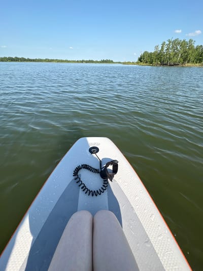 The image shows a point-of-view shot from a paddleboard on a body of water. In the foreground, the front of the paddleboard is visible, with a coiled leash and a person's legs. The water is calm with small ripples, and the background features a tree line along the horizon under a clear blue sky. The scene appears to be a natural setting, possibly a lake or a calm river, located in or near the city of Razdolnoye, Russia, given the context of the prompt.