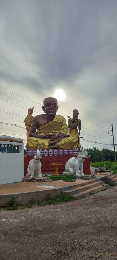 This image captures an outdoor sculpture in Ban Thung, Thailand. The sculpture depicts two monks in yellow robes, one seated and the other standing behind him, along with two white elephant statues. The sculpture is situated on a decorated platform with patterned backdrops. Overhead power lines stretch across the top of the frame, and vegetation is visible in the background. A section of a road or path borders the edge of the sculpture installation. The sky is overcast with the sun visible behind the clouds.