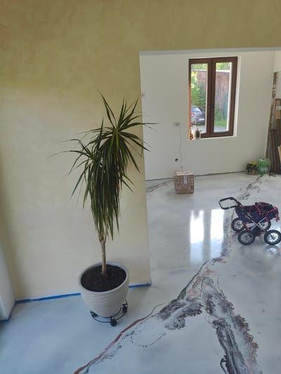 The image depicts an indoor room, seemingly in a state of recent renovation or furnishing, characterized by a modern and artistic aesthetic. The left wall exhibits a textured, light yellowish-beige finish, while the walls to the right and at the back are painted a clean white. The floor is a prominent feature, covered in a glossy, light grey or white epoxy resin with dramatic, abstract patterns of black, dark brown, and metallic copper-colored streaks, resembling flowing marble or geological formations.
In the foreground on the left, a tall Dracaena houseplant with long, spiky green leaves is potted in a grey, textured planter, which rests on a black wheeled caddy. Towards the right side of the reflective floor, a dark blue and pink child's stroller or doll's pram is visible.
A brown-framed window is centered in the back white wall, providing a view of a green outdoor area, a dark car with a European license plate, and a wooden fence, indicating a residential setting in Bad Pyrmont, Germany. On the windowsill below the window, a small potted plant and a brown glass bottle are placed. A cardboard box, bearing partially visible text "SOS S...", sits on the floor near the back wall. Electrical outlets are present on the white wall. In the far back right corner, construction materials, including wooden planks and transparent containers, are stacked. Blue painter's tape is visible along the base of the yellowish wall. Bright natural light streaming through the window suggests it is daytime. No individuals are present in the scene.